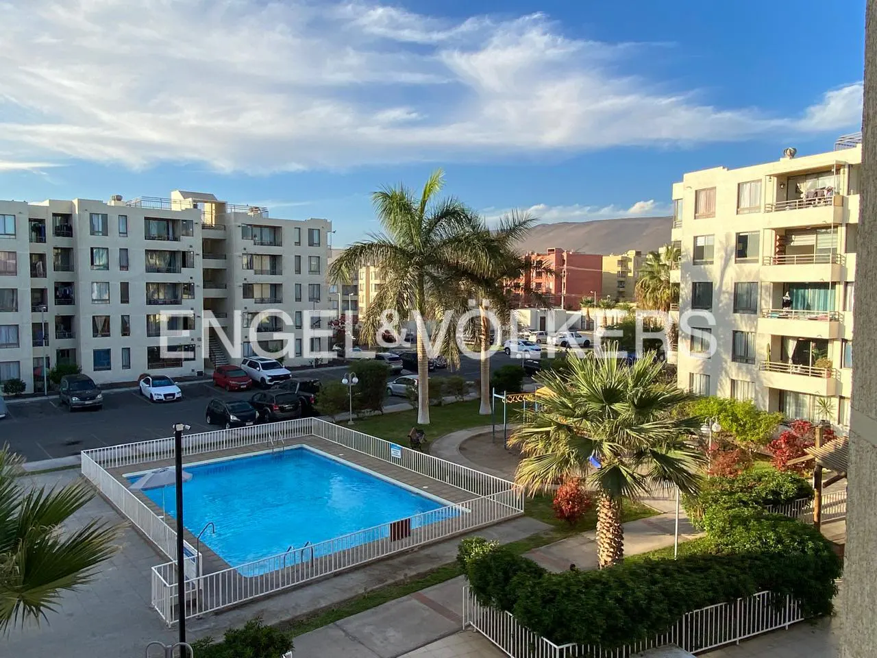 Aerial view of a complex with a pool, parking lot, and palm trees, under a blue sky with clouds. Engel & Völkers logo is superimposed.