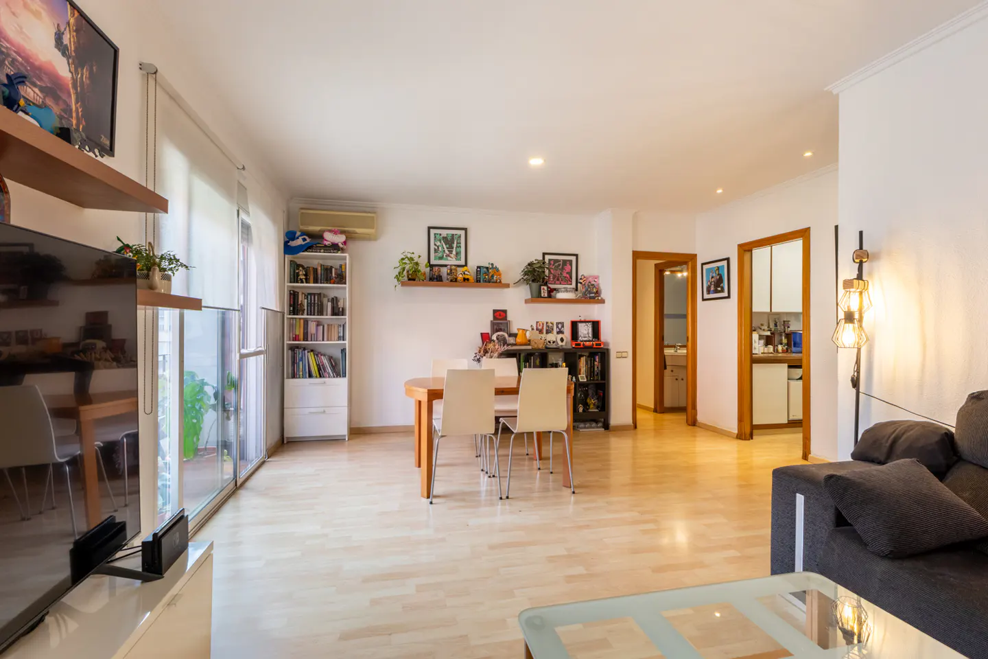 Bright living room with wood floors, dining table with four chairs, and a gray sofa. Shelves and decorations add personality.