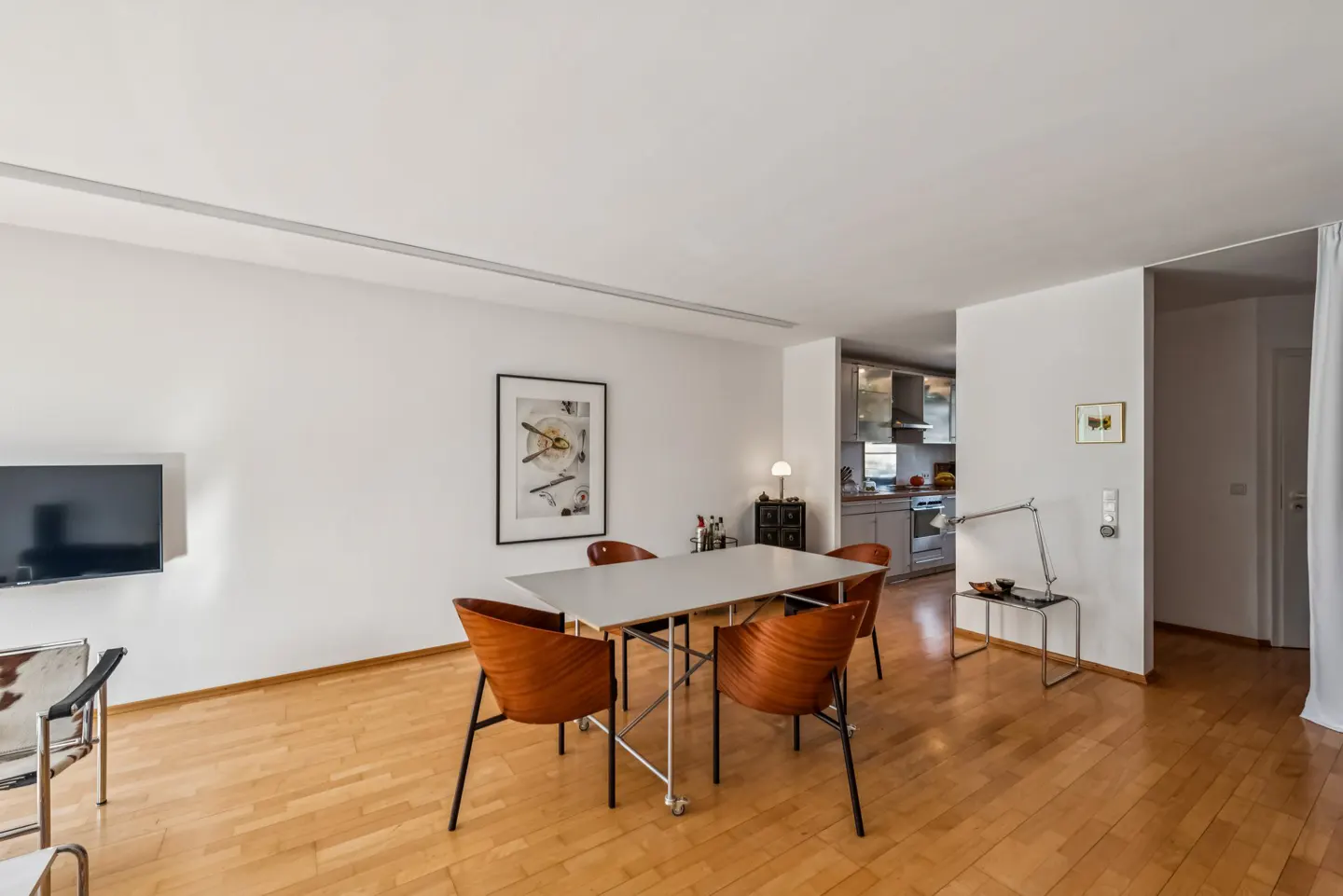Bright living room with wood floors, white walls, and a view into the kitchen. A white table with four wood chairs sits in the center. A TV hangs on the wall.