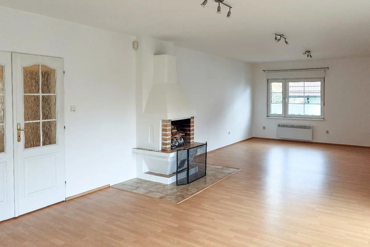Bright, empty living room with wood floors, white walls, and a white fireplace with a black screen. Double doors to the left and a window in the back.