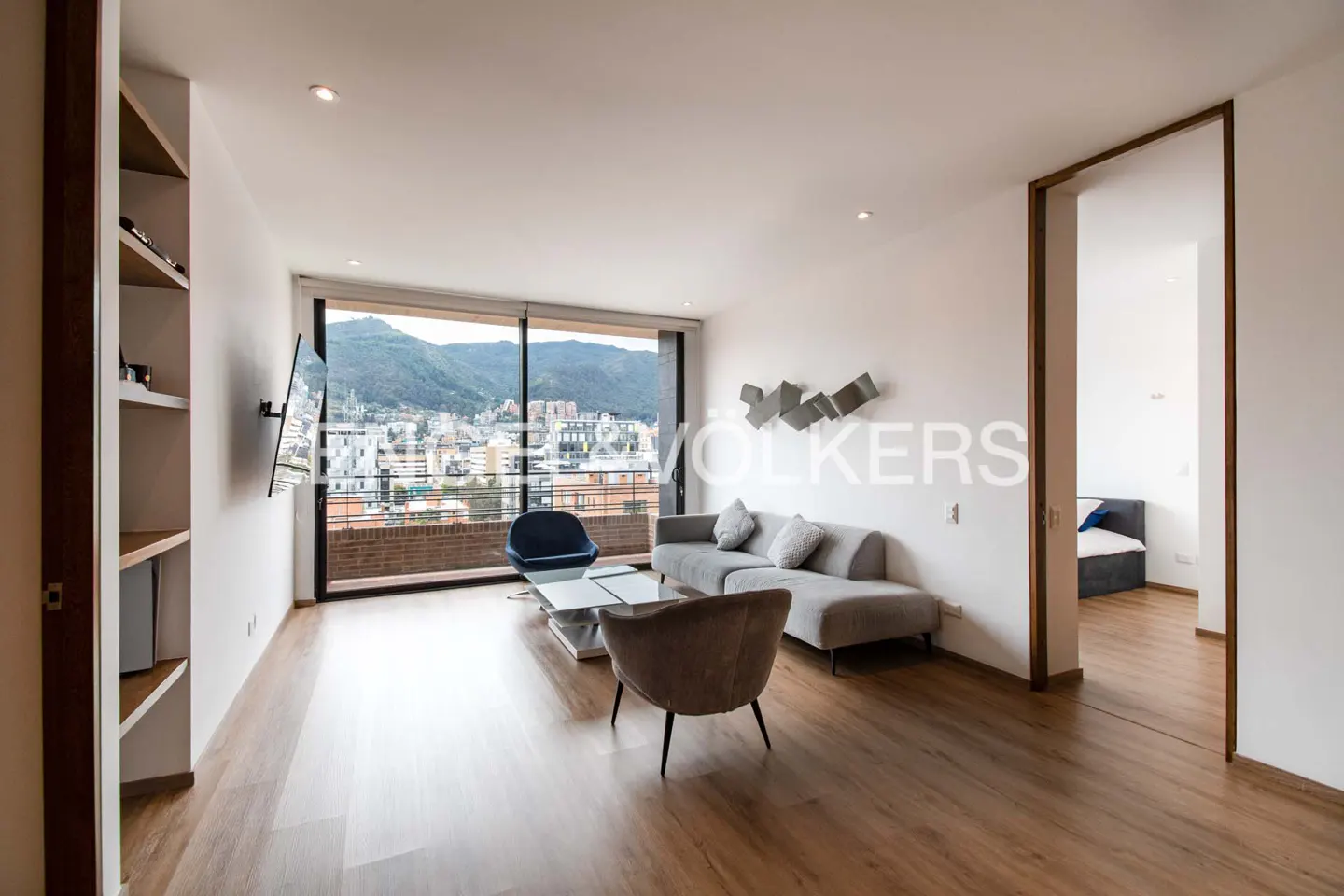 Bright living room with wood floors, gray sofa, blue chair, and city view through large windows. Bedroom visible through doorway.