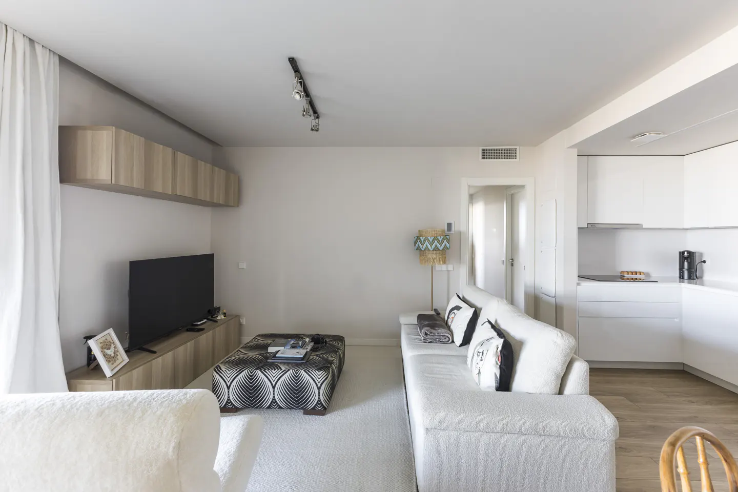 Bright living room with white sofa, patterned ottoman, TV, and open kitchen. Light wood floors and cabinets add warmth.