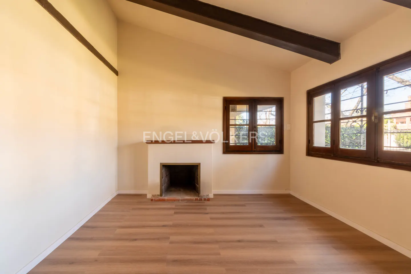 Empty room with wood floors, fireplace, and two wood-framed windows. Dark wood beams on the ceiling. Cream-colored walls.