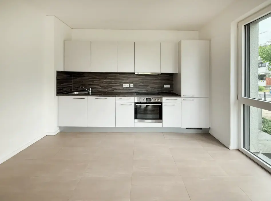 Modern kitchen with white cabinets, dark horizontal backsplash, stainless steel oven, and light tile floors near a window.