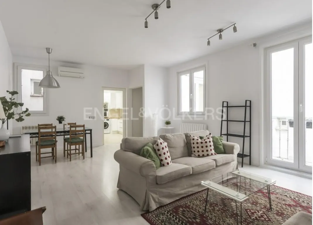 Bright, open living space with white walls, light wood floors, and a beige sofa with colorful pillows. Dining table and kitchen visible.