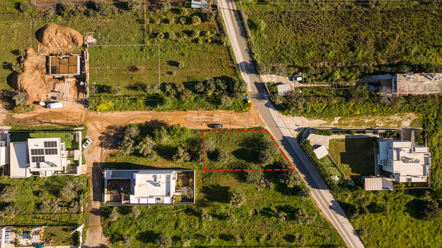 Aerial view of a vacant lot outlined in red, surrounded by green grass, trees, and houses. A road runs along one side of the property.
