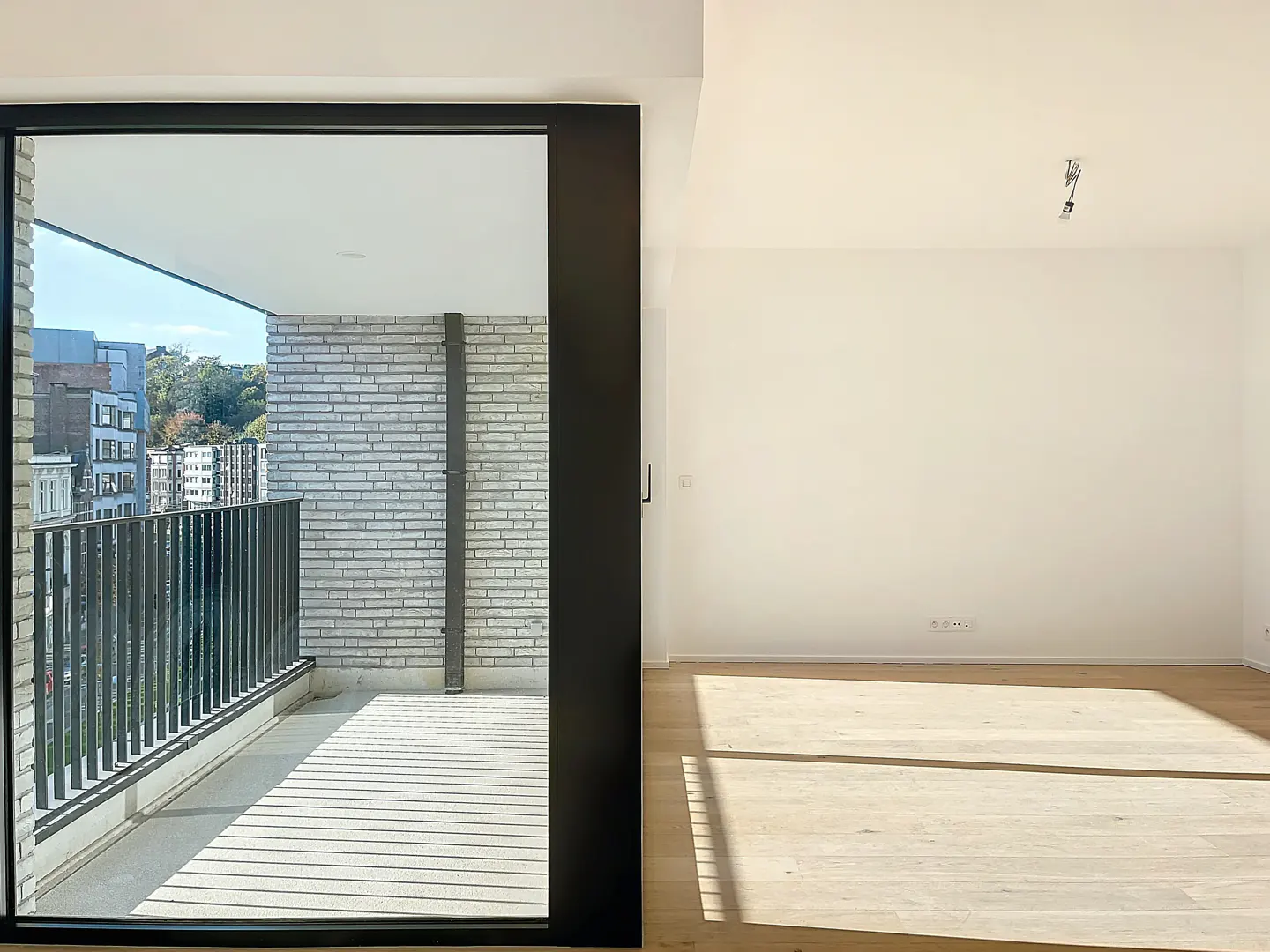 A bright, empty room with light wood floors and white walls. A sliding glass door leads to a balcony with a gray brick wall and black railing.