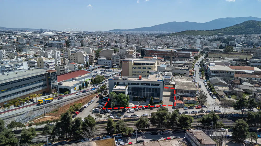 Aerial view of a city with buildings, cars, and mountains in the background. A red dotted line outlines a property.