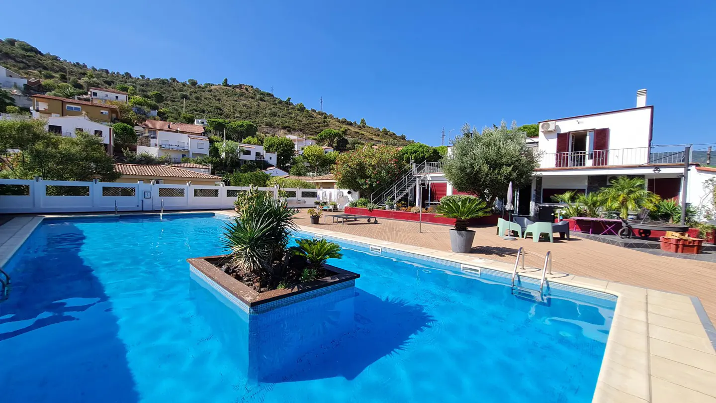 A bright blue swimming pool with a small island of plants, a wooden deck, and a white house under a clear blue sky.