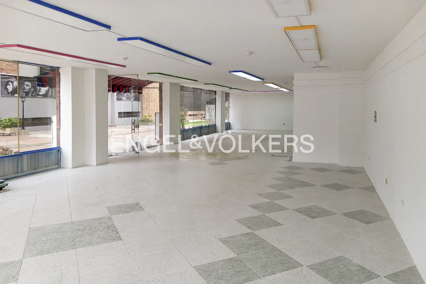 Empty commercial space with white walls, gray tile floor, and colorful ceiling lights. Glass doors lead to an outdoor area.