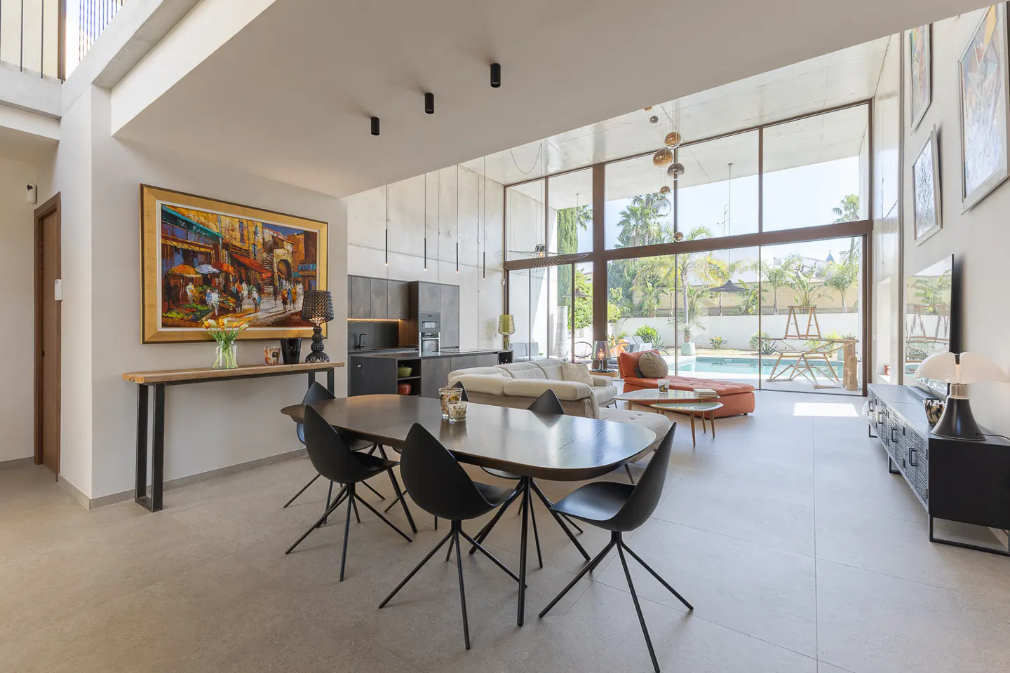 Open-concept living space with dining table, black chairs, and large windows overlooking a pool and lush greenery. Artwork adorns the walls.