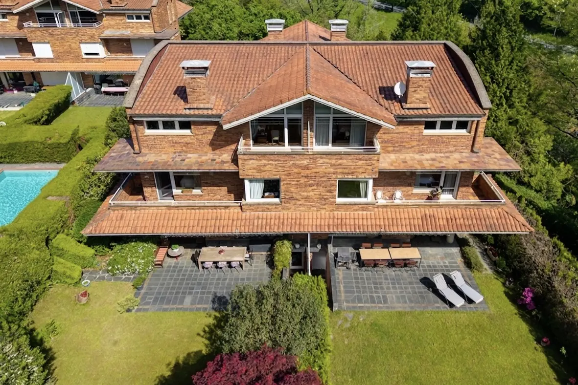 Aerial view of a brick house with a red tile roof, a patio with tables and chairs, and a green lawn.