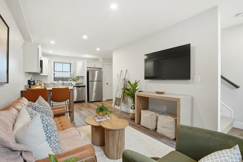 A bright, open-concept living space with a brown leather sofa, wood coffee tables, and a kitchen in the background.