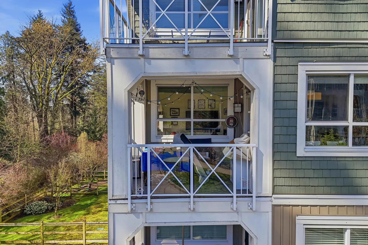 Exterior view of a three-story building with white balconies and green siding, overlooking a grassy yard and trees.