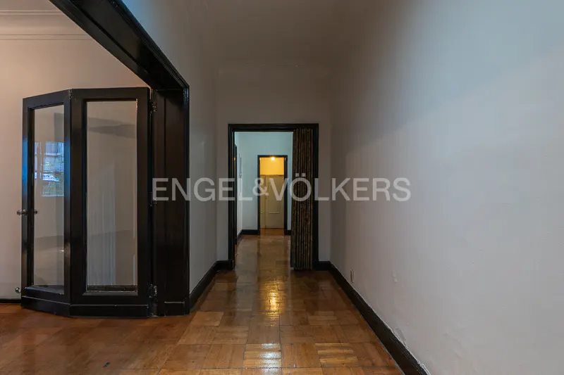Interior view of a hallway with parquet floors, white walls, and black trim. A glass door is on the left, and a doorway leads to a brightly lit room.