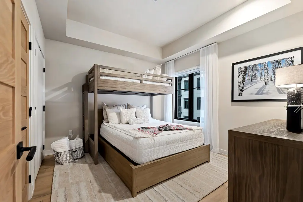 Bedroom with a light wood bunk bed, white bedding, and a window with sheer white curtains. A dresser and framed winter scene art are on the right.