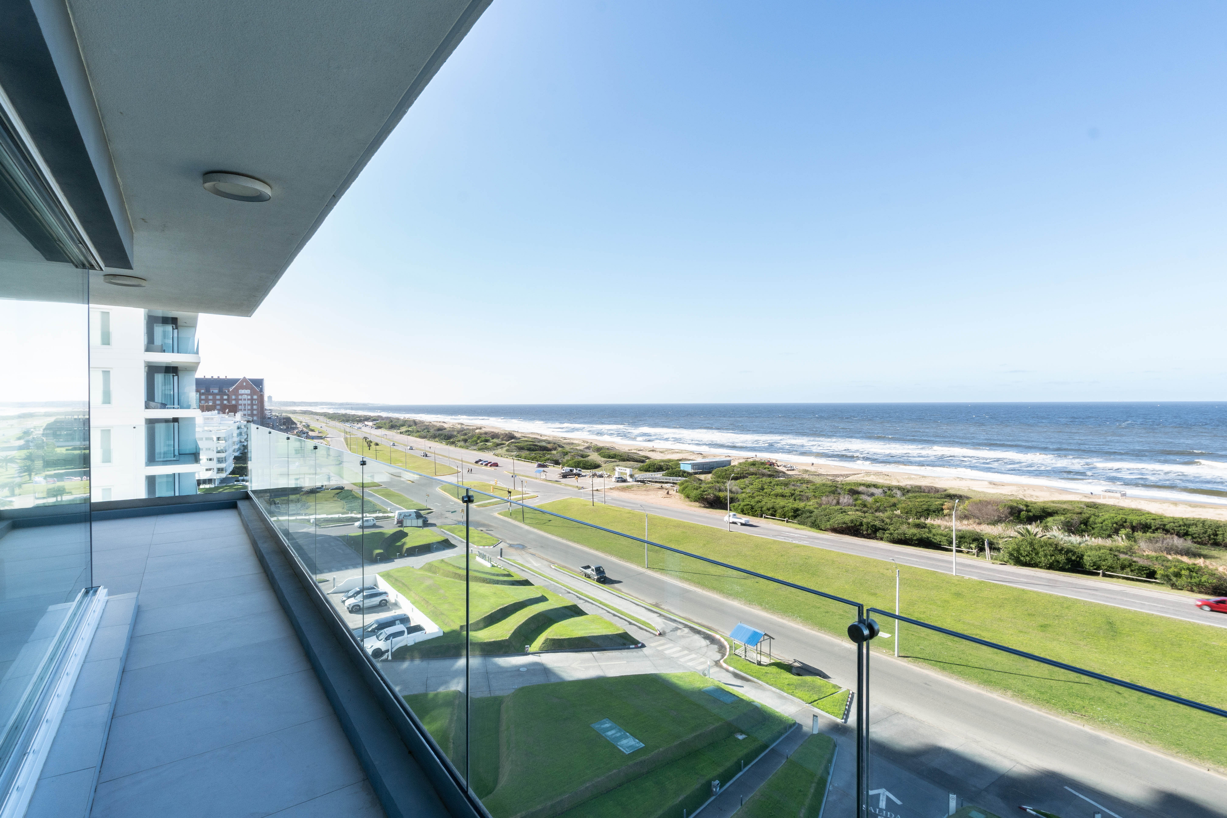 View from a modern balcony overlooking a coastal road, green lawns, and the blue ocean under a clear sky.