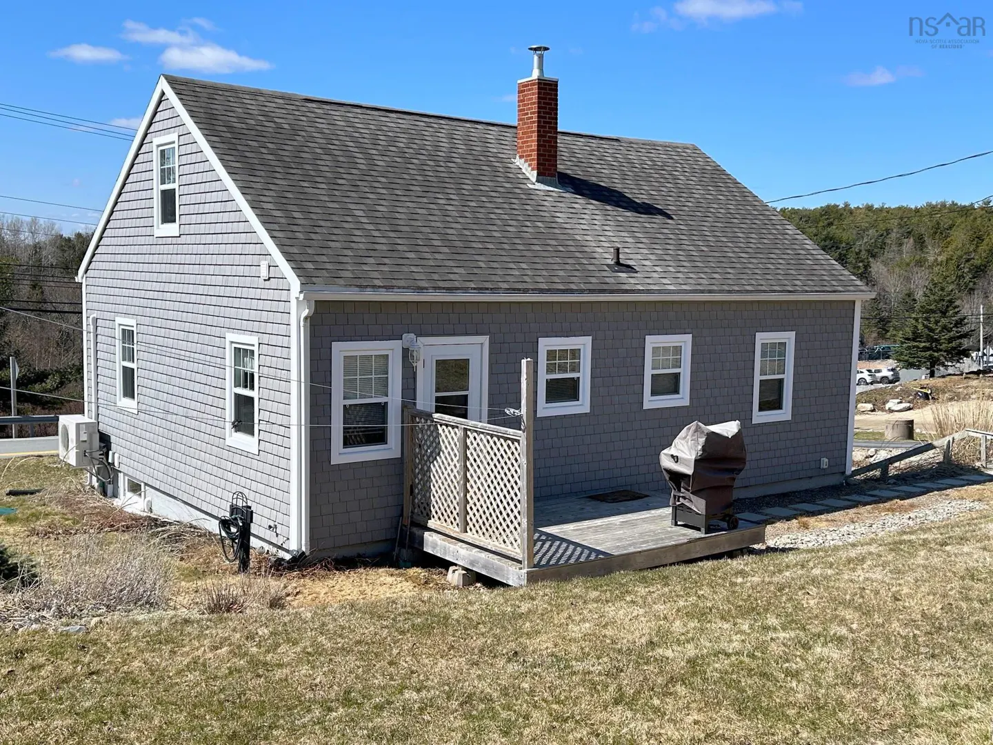 Exterior view of a gray shingle house with a gray roof, white trim, and a red brick chimney on a sunny day. A wooden deck and grill are visible.