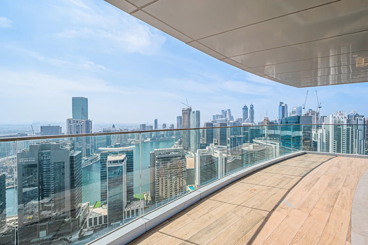 Balcony view of a city skyline. The balcony has a wood floor and glass railing. Tall buildings and a blue sky are in the background.