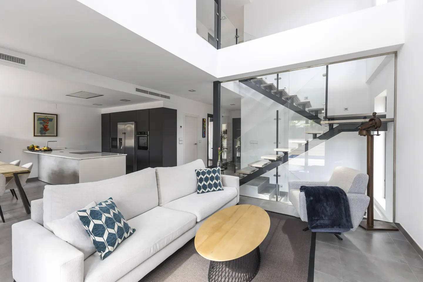 Bright, modern living room with white sofa, wood coffee table, and glass-enclosed staircase. Kitchen island and dining area visible in the background.