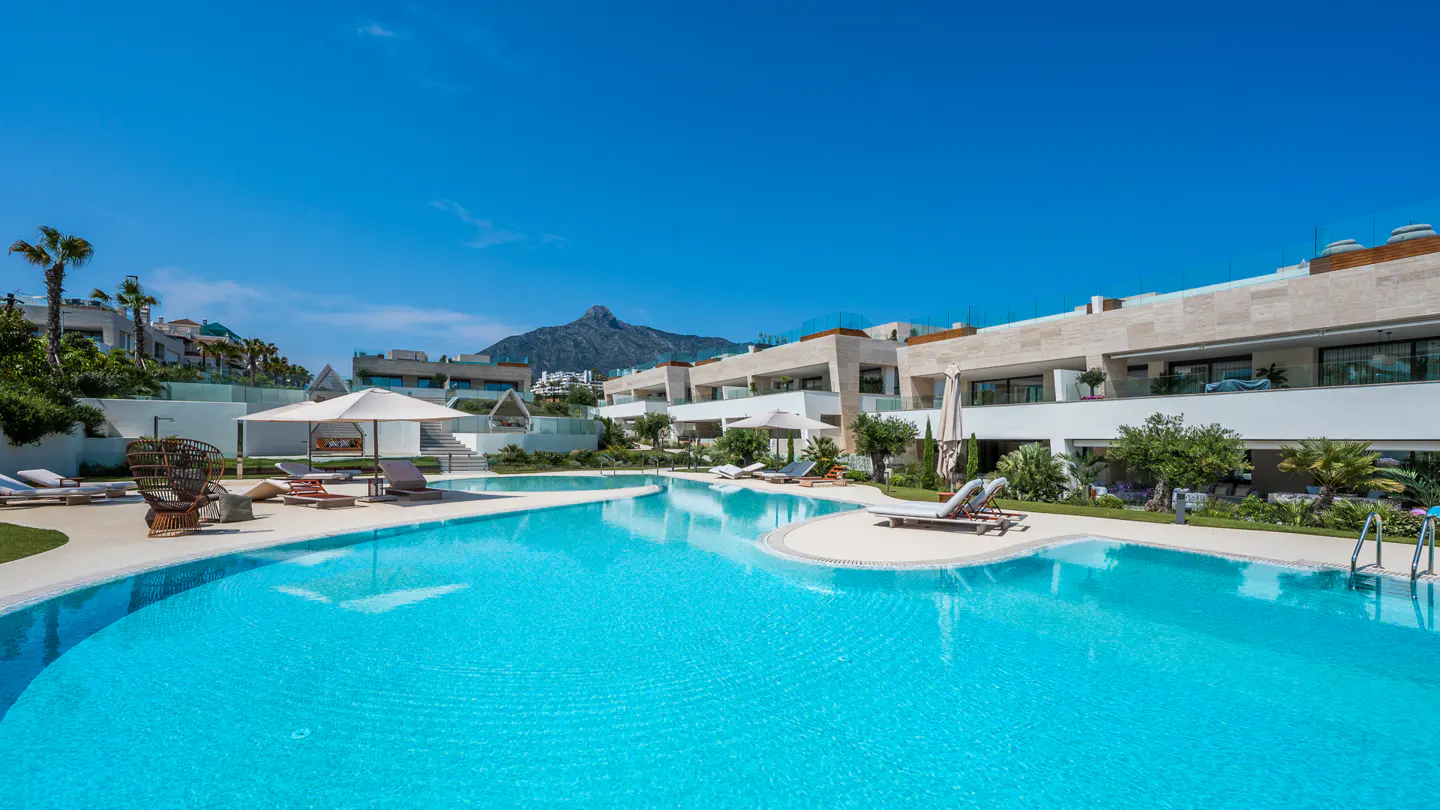 Luxury resort with a turquoise pool, lounge chairs, and modern buildings under a clear blue sky. Mountain in the background.