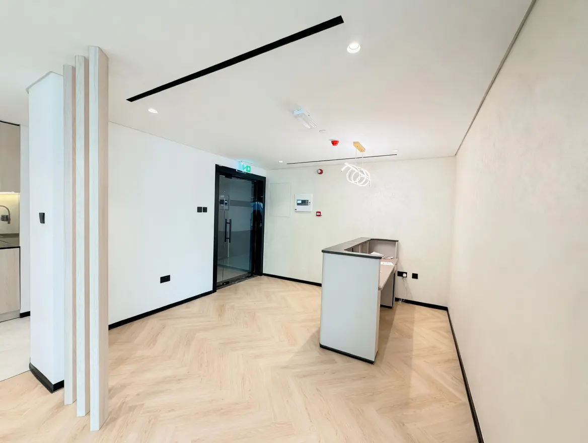 A modern office space with a reception desk, glass door, and herringbone wood floors. The walls are white, and the ceiling has black accents.