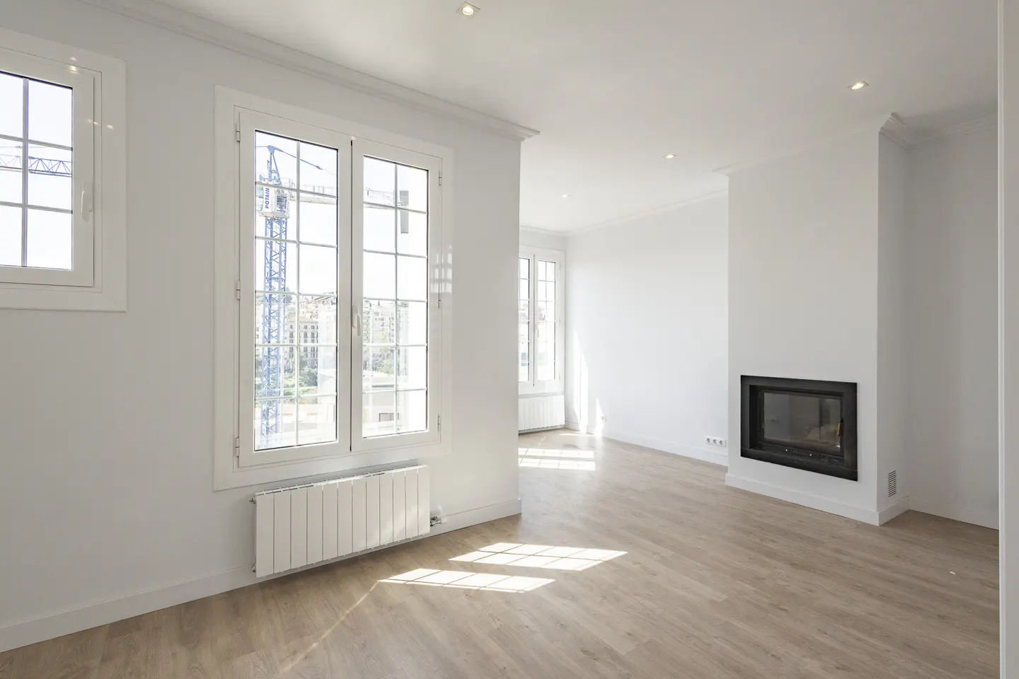 Bright, empty room with light wood floors, white walls, and a black fireplace. Windows show a city view with a construction crane.
