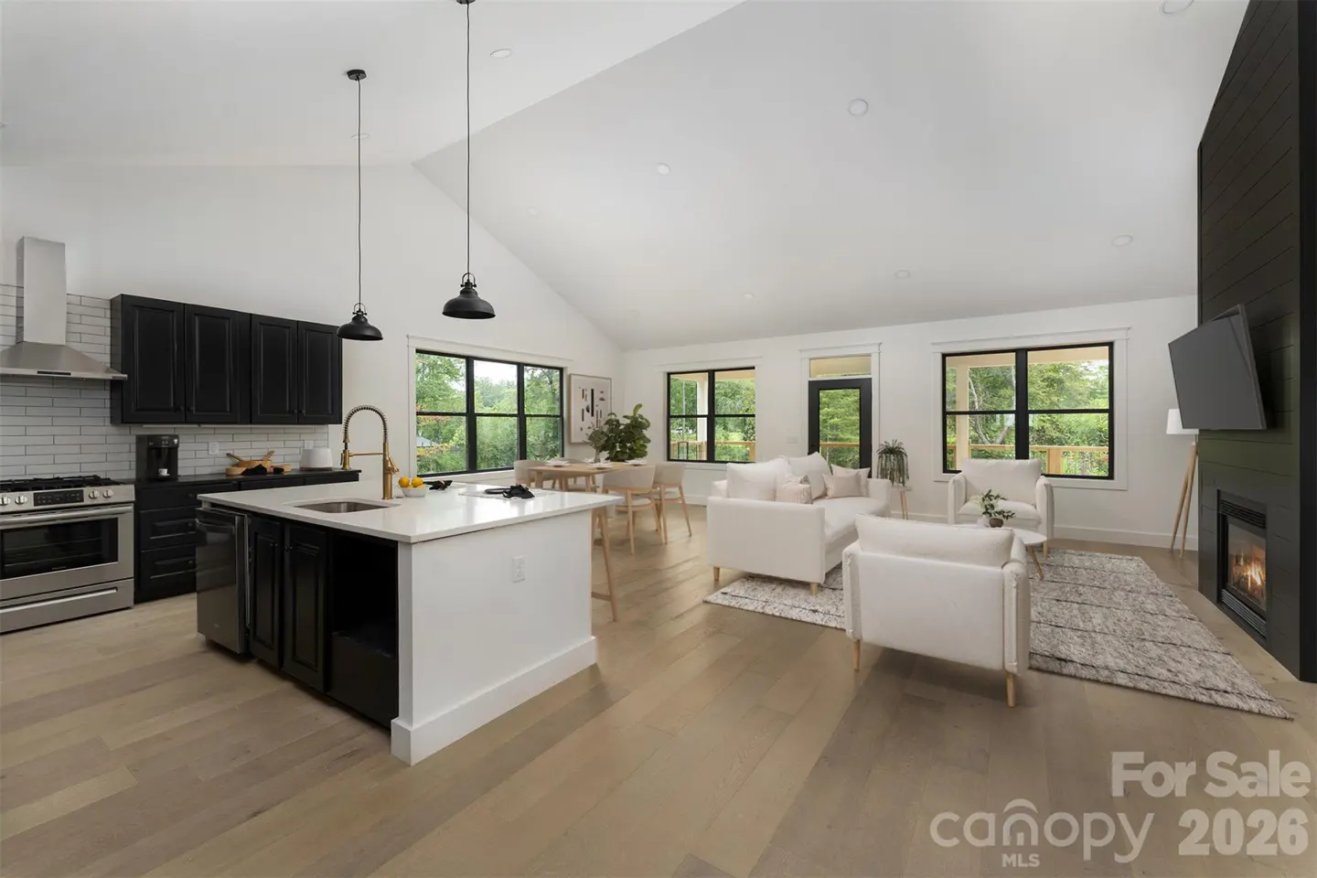 Open-concept living space with a kitchen island, white sofas, and a fireplace. Black cabinets and window frames add contrast.