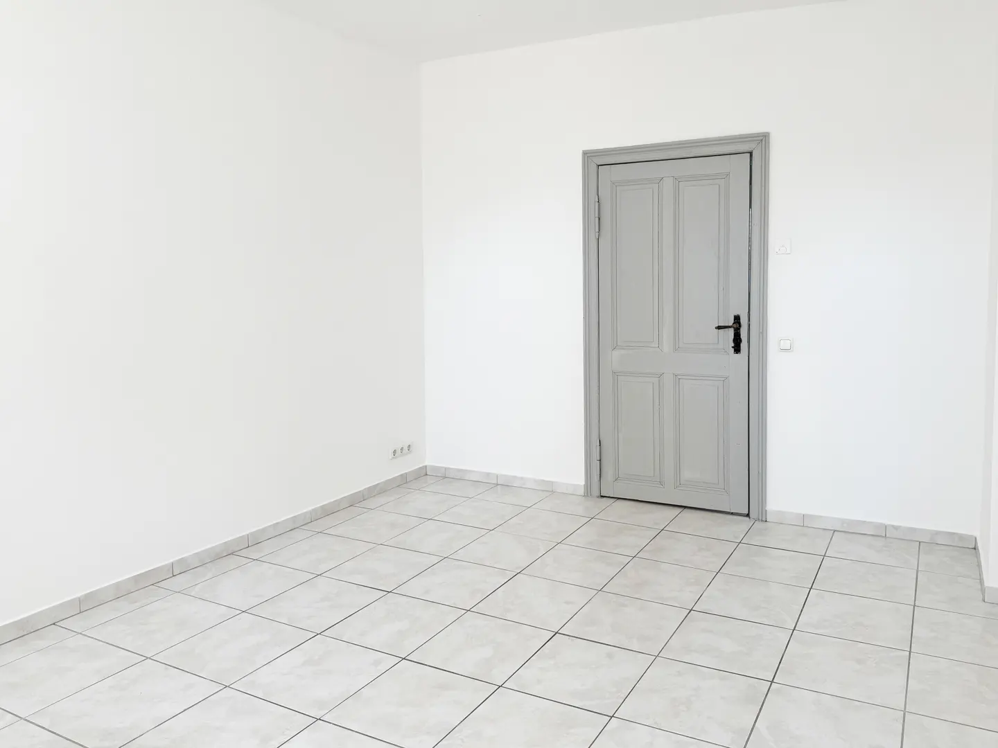 Empty room with white walls, light gray tile floor, and a gray door with a black handle.
