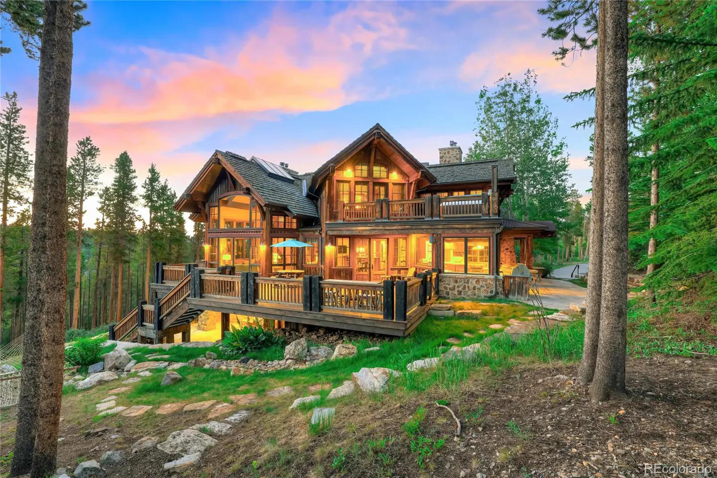 Exterior view of a large, multi-story wood and stone house with a deck, surrounded by trees and a colorful sunset sky.