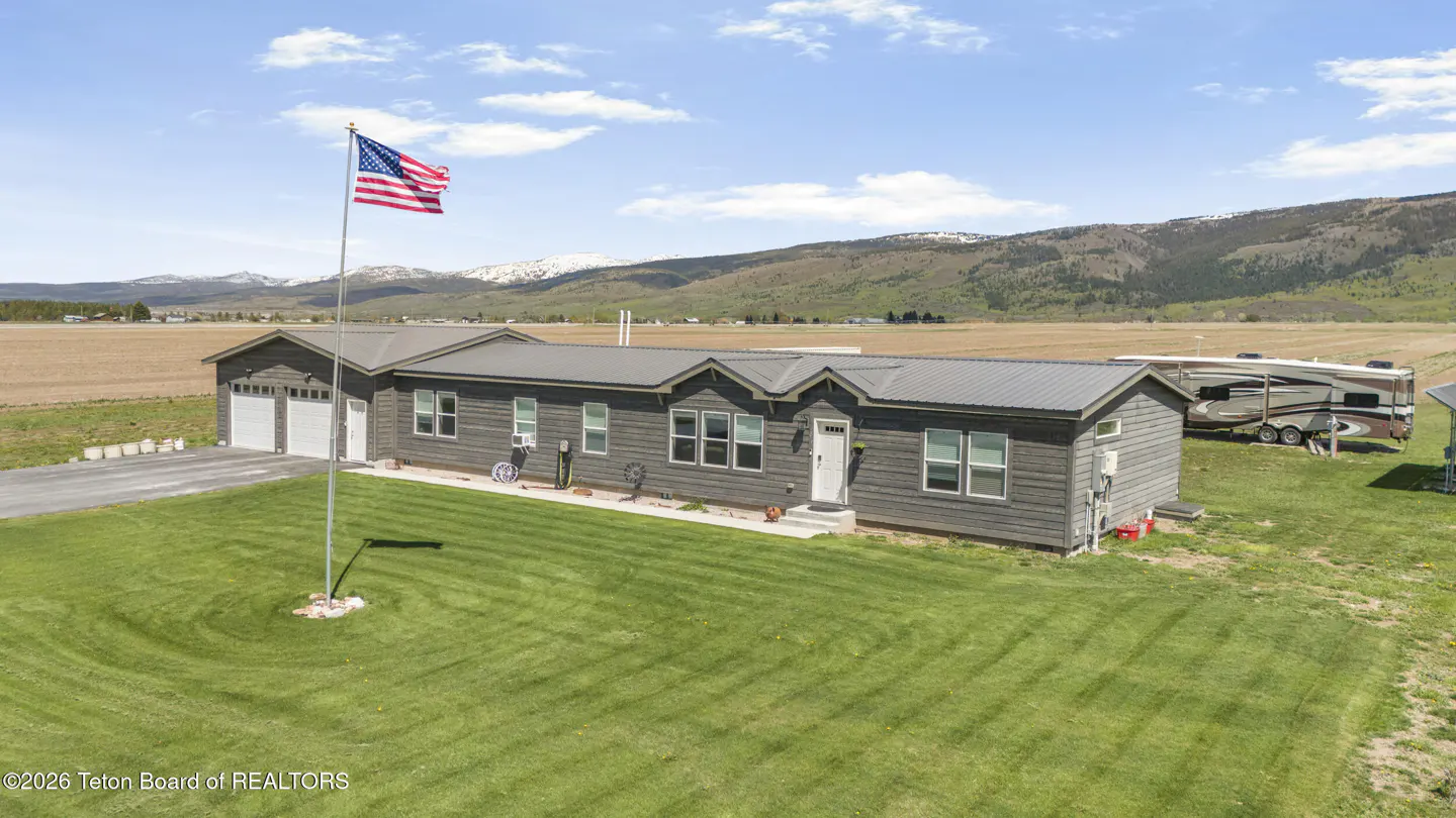 Exterior view of a gray house with a metal roof, a green lawn, and an American flag in front of a mountain range.