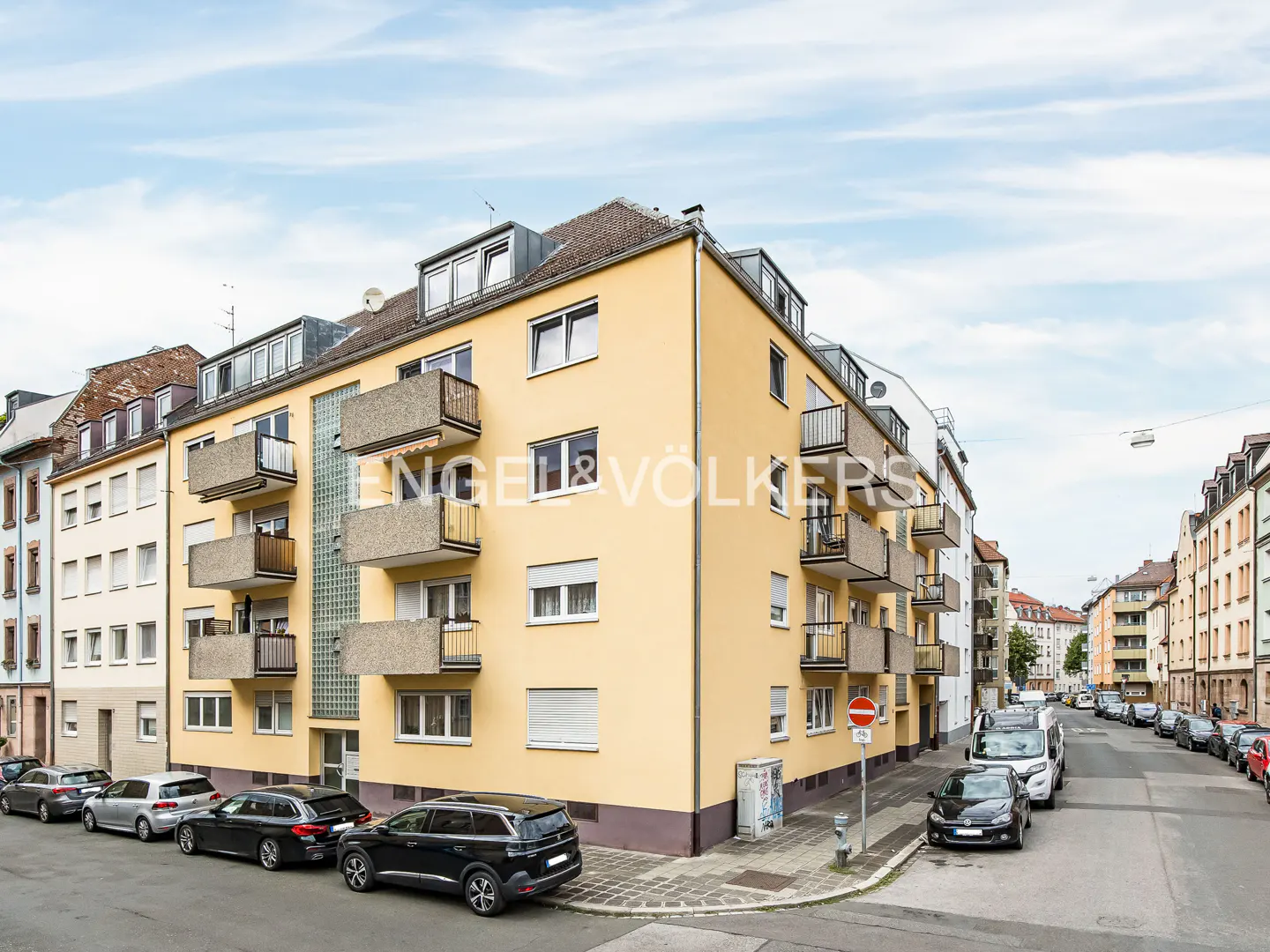 A yellow multi-story apartment building with concrete balconies on a city street with parked cars and a blue sky.