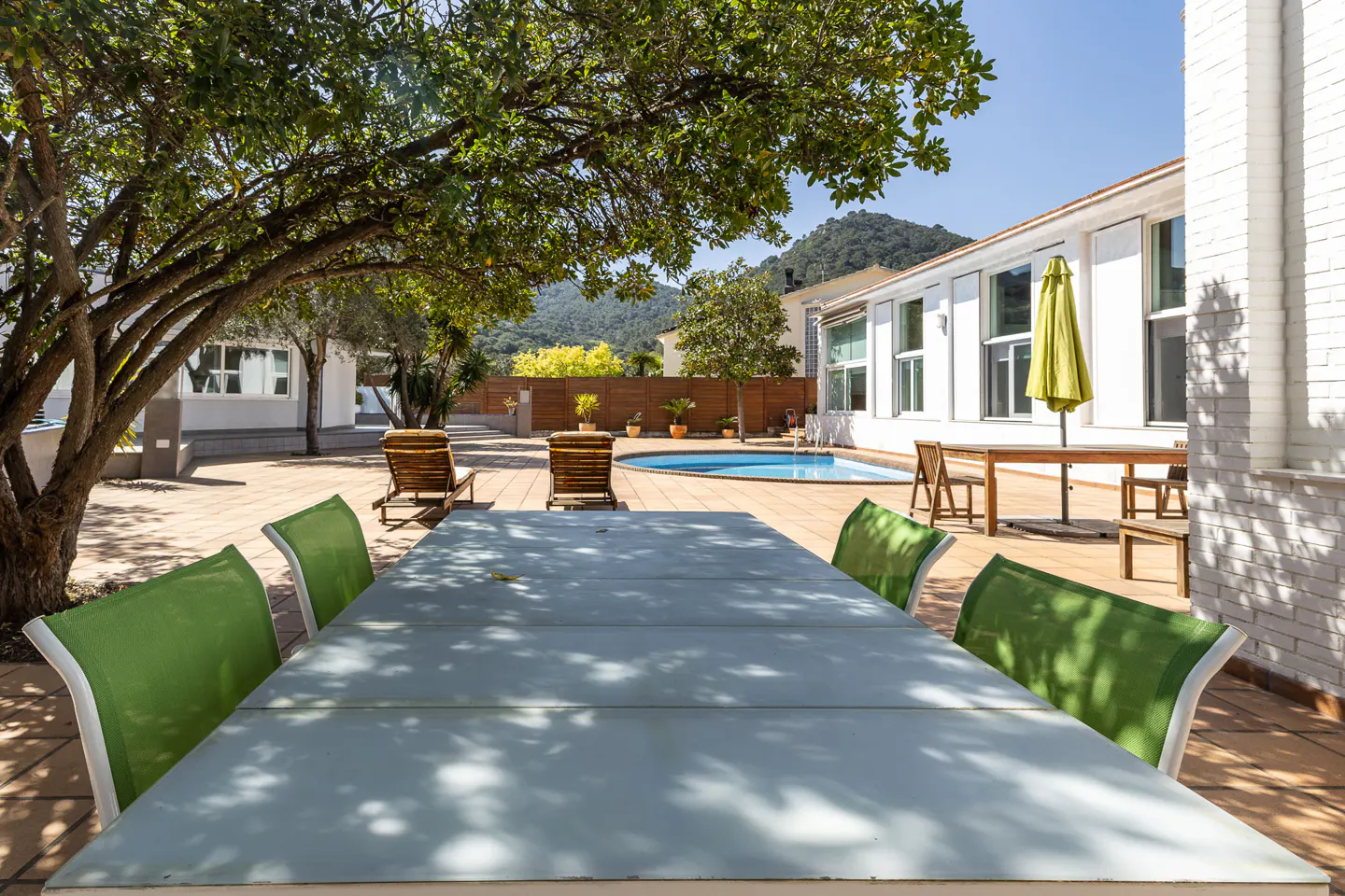 Outdoor patio with a large table, green chairs, pool, lounge chairs, and a white building in the background.