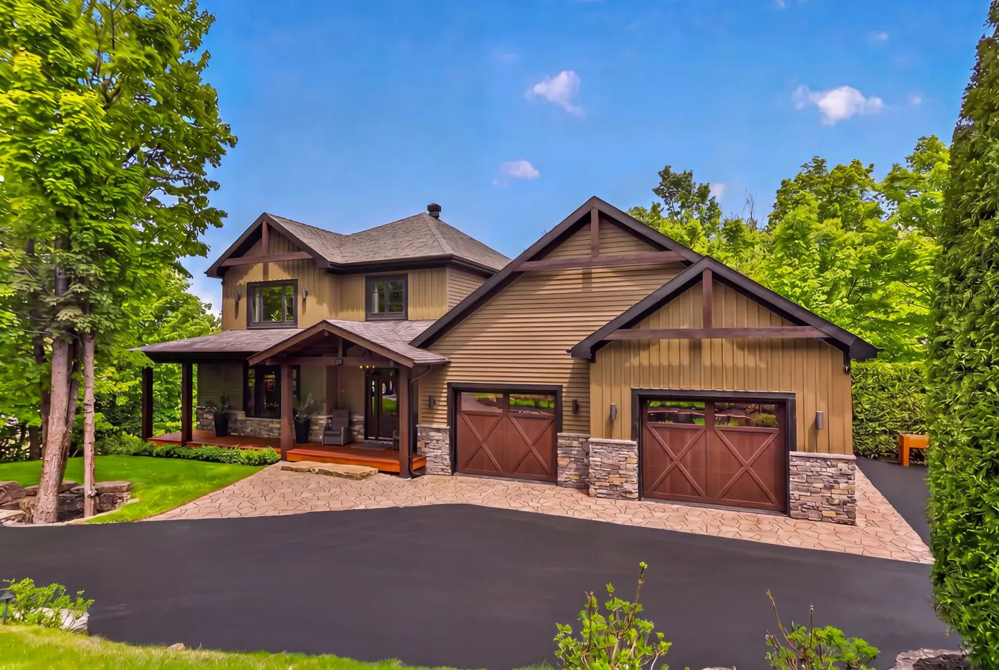 Two-story home with olive siding, stone accents, and a two-car garage, set on a paved driveway surrounded by lush trees.