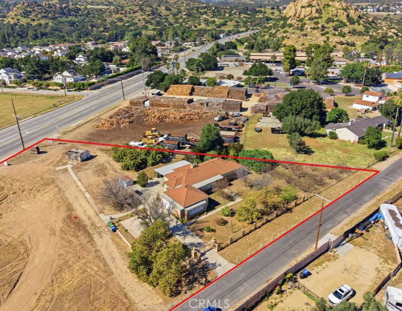 Aerial view of a property with a red boundary line, featuring a house with a red tile roof, trees, and a road. In the background, there are piles of wood and a mountain.