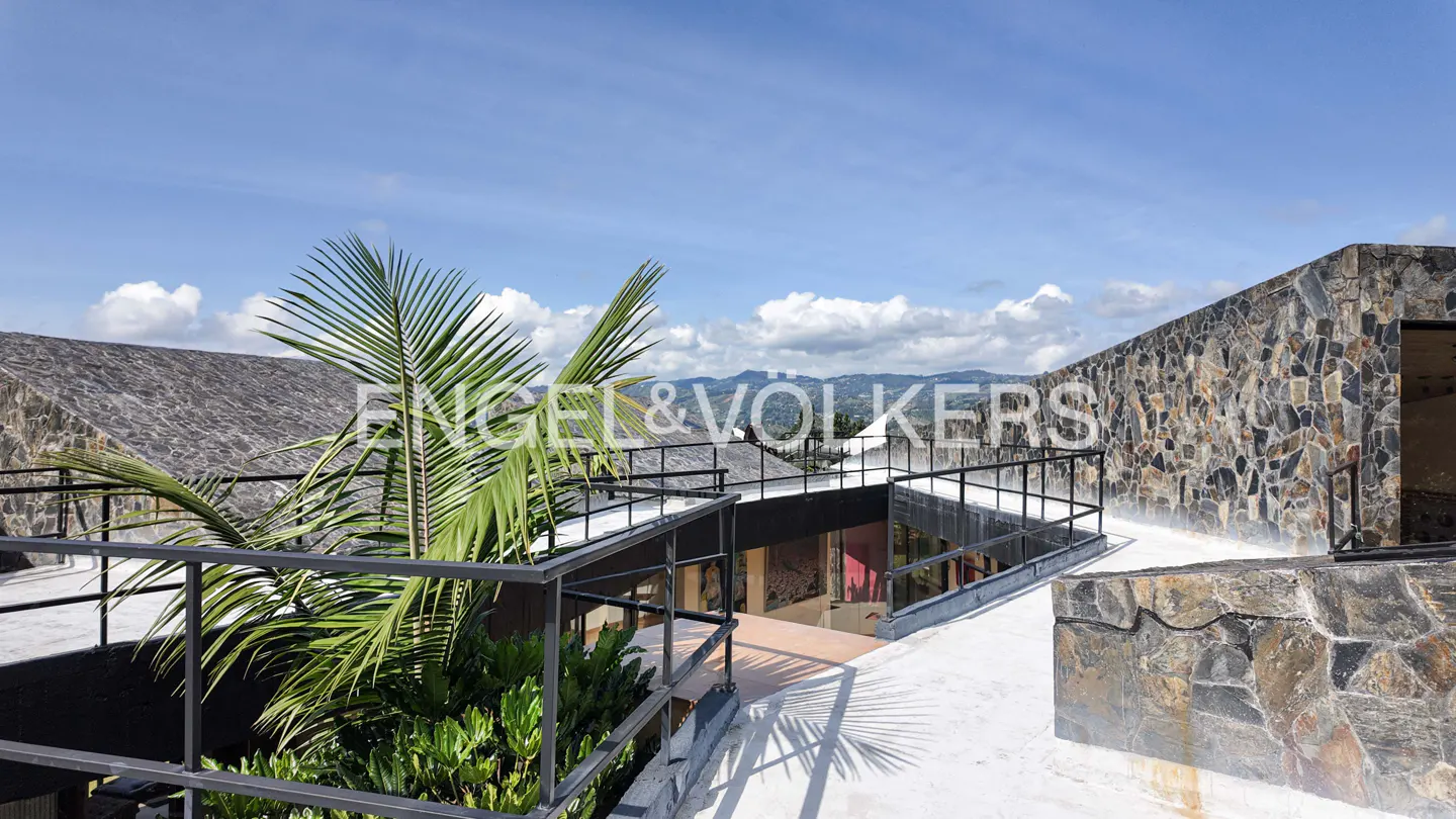 Rooftop view of a stone house with black railings, palm tree, and mountain view under a blue sky with clouds.