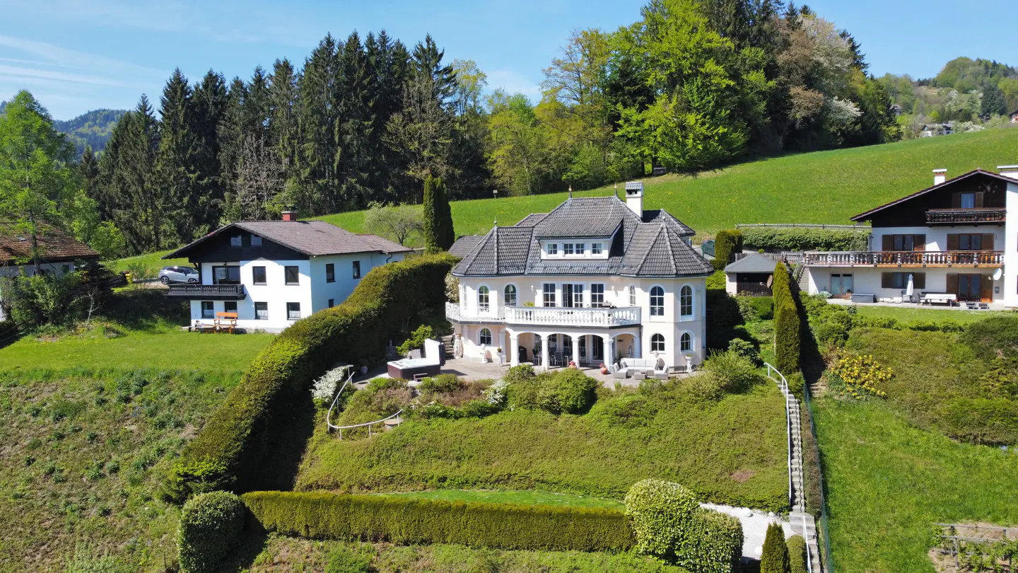 A two-story cream house with a gray roof sits on a green hillside with trees in the background.