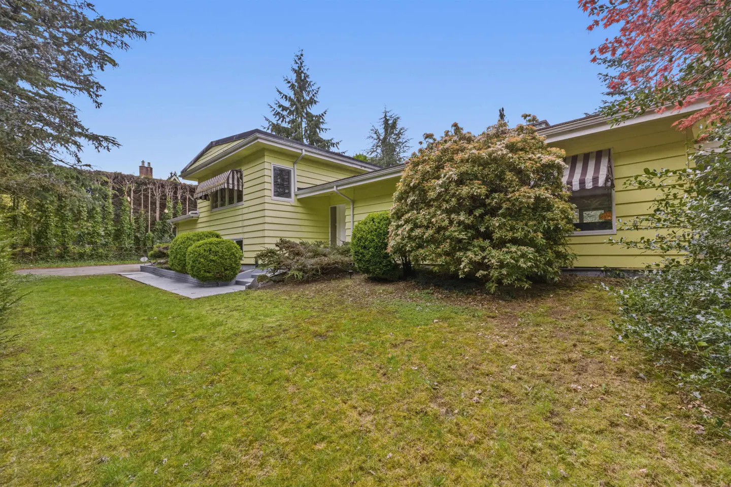 A yellow, two-story house with black and white striped awnings sits on a green lawn under a blue sky.