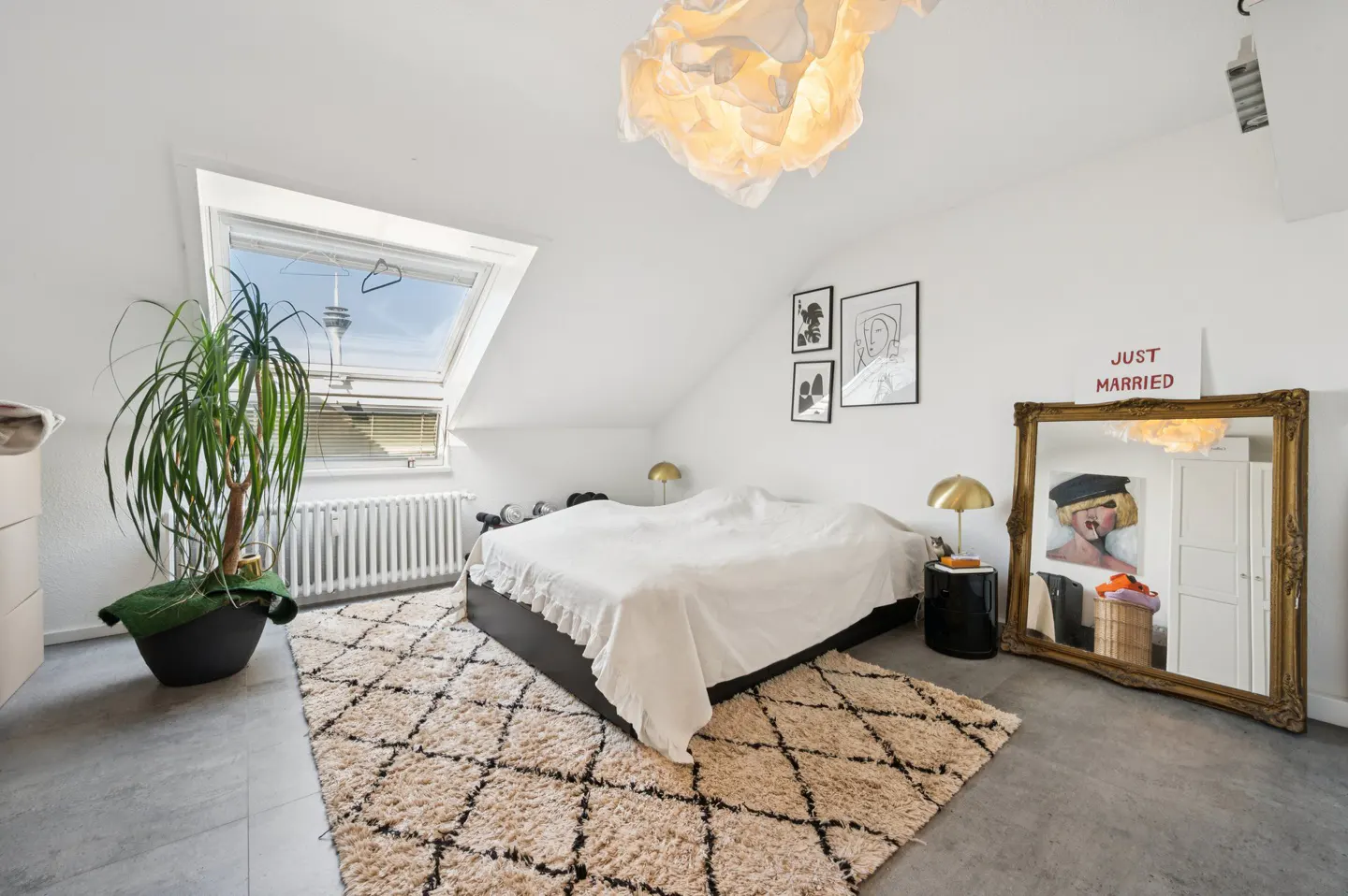 Attic bedroom with white walls, a bed, a patterned rug, and a large gold-framed mirror with a "Just Married" sign. A skylight shows a tower.