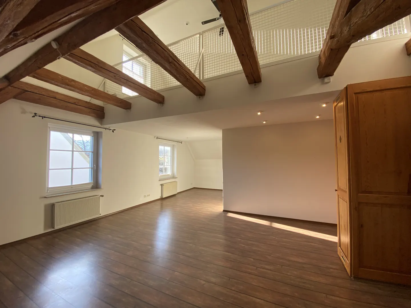 Spacious attic room with dark wood floors, white walls, exposed wooden ceiling beams, and a loft area with a metal railing.