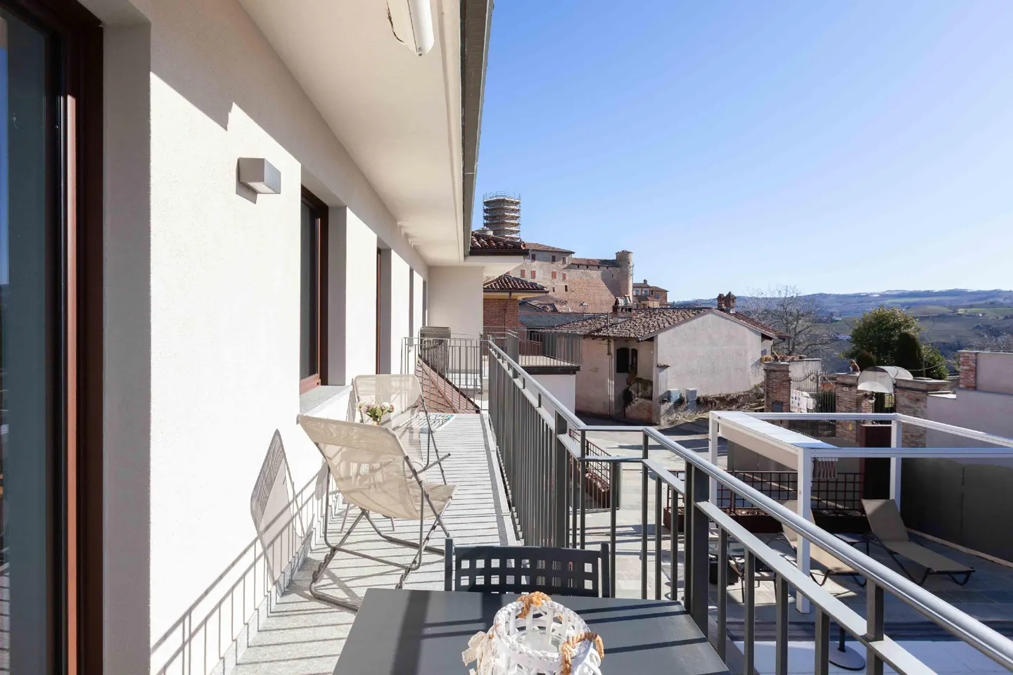 A bright balcony with gray metal railings overlooks a European town with a castle in the background. A table and chairs sit on the balcony.