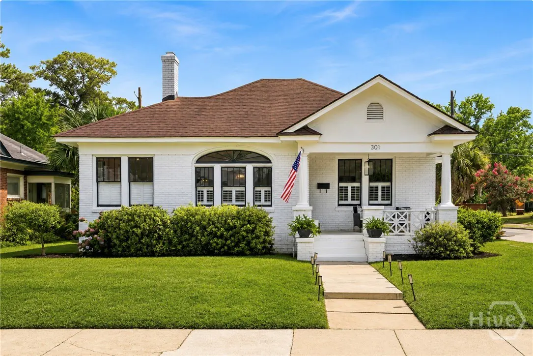 Charming white brick bungalow with a brown roof, front porch, American flag, and manicured lawn under a blue sky.