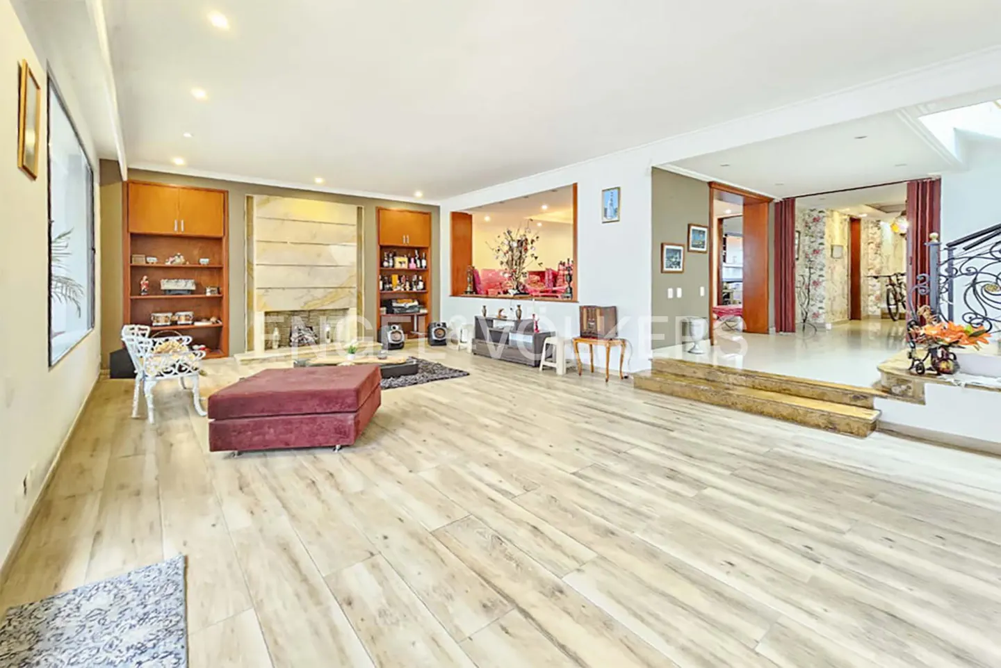 Wide shot of a living room with light wood floors, a red ottoman, and built-in shelves flanking a fireplace.