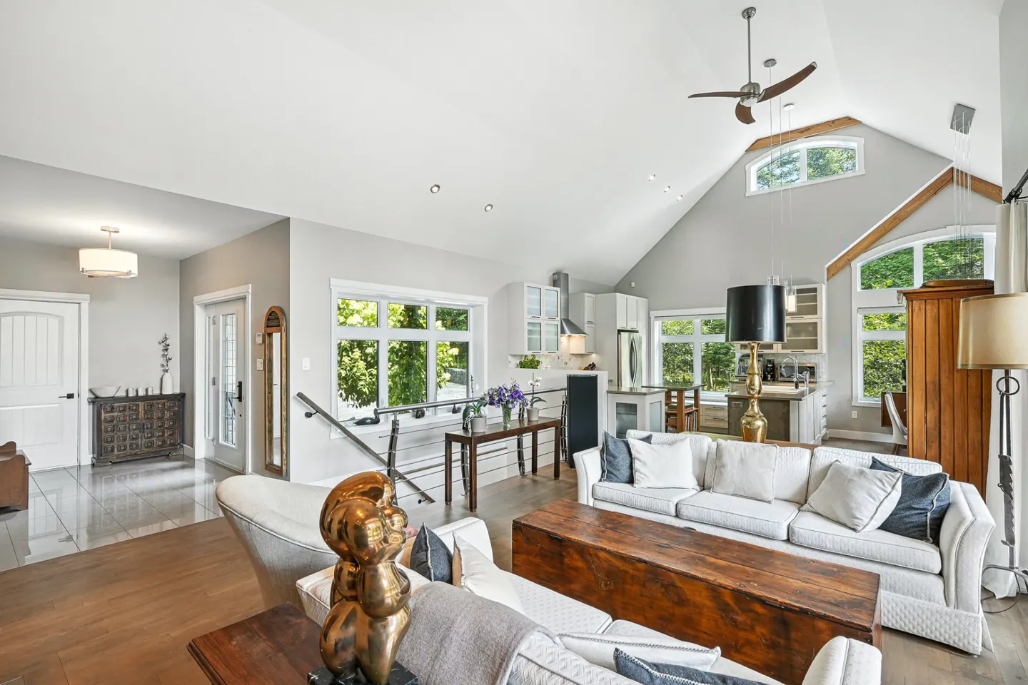 Bright, open-concept living room with white sofas, wood floors, and a vaulted ceiling. Kitchen visible in the background with white cabinets.