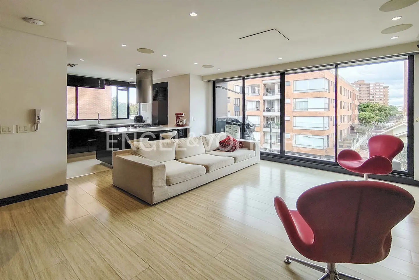 Open-concept living space with a white sofa, red chairs, and a kitchen island. Large windows offer a city view.