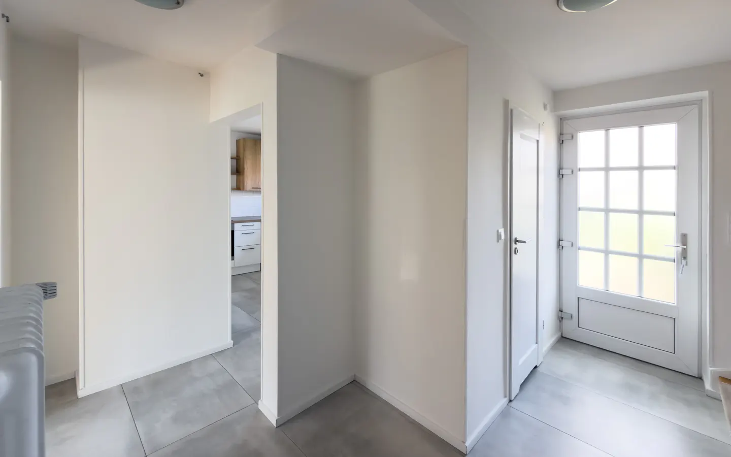 Bright, white hallway with gray tile floor. A doorway leads to a kitchen with light wood cabinets. A white front door with glass panes is on the right.