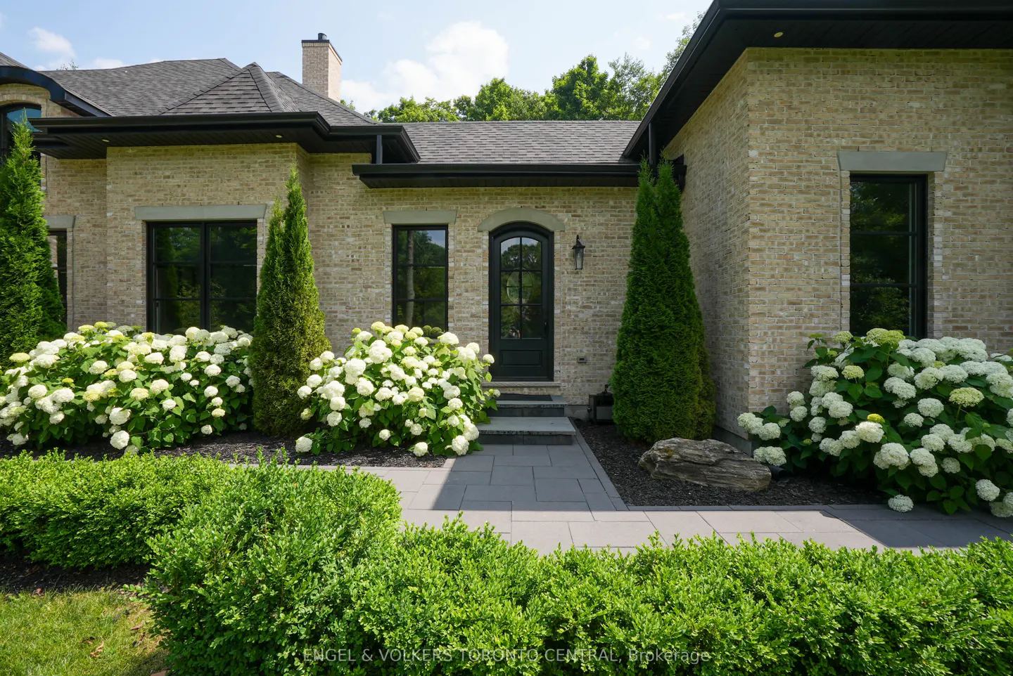 Front exterior of a tan brick house with a black door, black windows, and white hydrangeas.