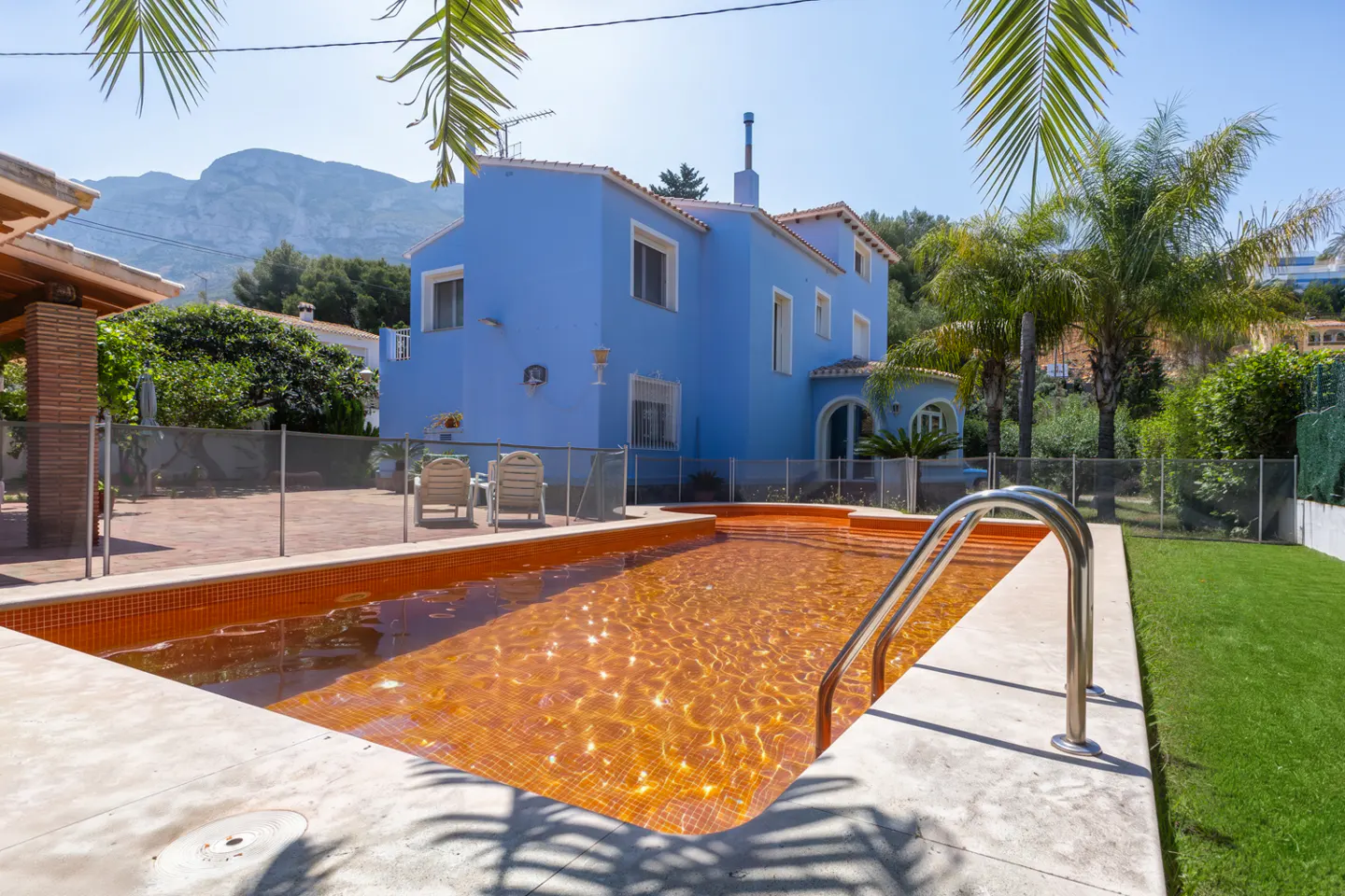 Bright blue house with a tiled swimming pool, patio, and palm trees against a mountain backdrop on a sunny day.