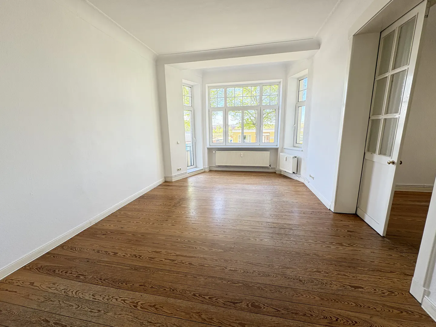 Empty room with hardwood floors, white walls, and a bay window with a radiator. A white door is partially open.