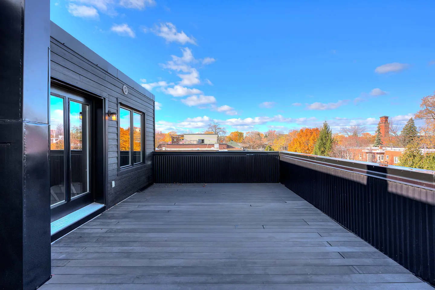 Black wood deck with a black metal railing, door and window. Fall foliage and blue sky in the background.
